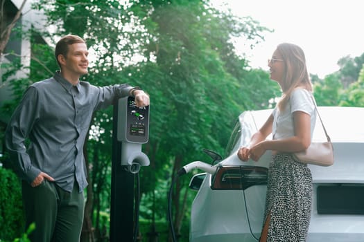 Young couple travel with EV electric car charging in green sustainable city outdoor garden in summer shows urban sustainability lifestyle by green clean rechargeable energy of electric vehicle innards