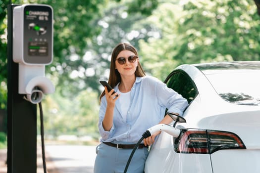 Young woman using smartphone online banking application to pay for electric car battery charging from EV charging station during vacation holiday road trip at national park or summer forest. Exalt