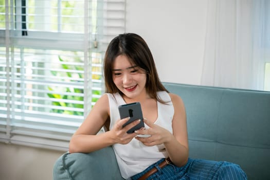 Social media networks. Happy beautiful female relax and shopping online with smartphone, Smiling Asian woman sitting on sofa reading news on mobile smart phone at living room at home