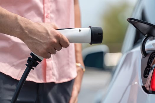 Hand insert EV charger plug into electric vehicle to recharge EV car battery from outdoor charging station. Fresh daylight environment with alternative clean and sustainable energy concept. Perpetual