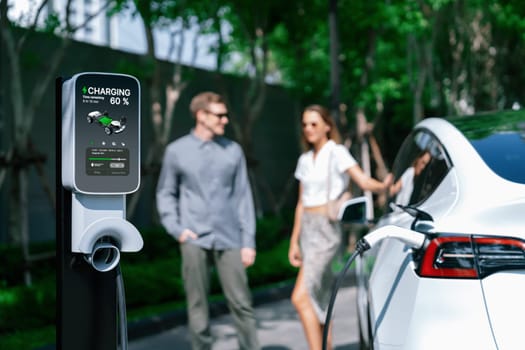 Young couple travel with EV electric car charging in green sustainable city outdoor garden in summer shows urban sustainability lifestyle by green clean rechargeable energy of electric vehicle innards