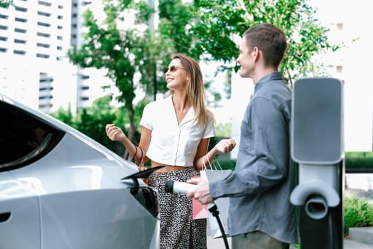 Young couple travel with EV electric car charging in green sustainable city outdoor garden in summer shows urban sustainability lifestyle by green clean rechargeable energy of electric vehicle innards