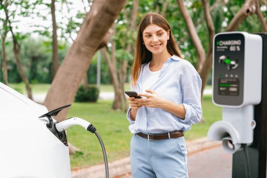 Young woman using smartphone online banking application to pay for electric car battery charging from EV charging station during vacation holiday road trip at national park or summer forest. Exalt