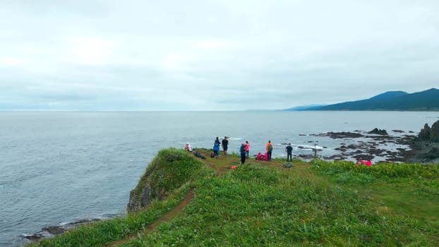 Top view of group of tourists on edge of sea cliff. Clip. Hikers on edge of rocky coastline. Tourists on edge of cliff of seashore on cloudy day.