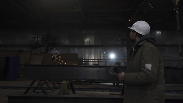 Man walks in helmet at factory on background of workers. Clip. Man looks at work of factory workers. Man walks on background of worker with sparkling welding.