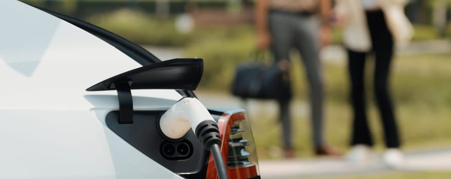 Young couple recharge electric car battery from charging station in green city park in springtime. Rechargeable EV car for sustainable environmental friendly urban travel lifestyle. Panorama Expedient