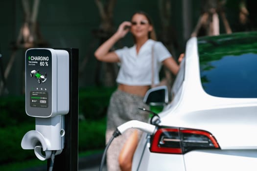 Young woman travel with EV electric car charging in green sustainable city outdoor garden in summer. Urban sustainability lifestyle by green clean rechargeable energy of electric BEV vehicle innards