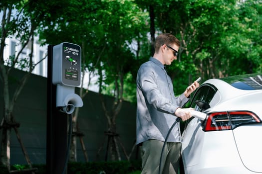 Young man travel with EV electric car charging in green sustainable city outdoor garden in summer shows urban sustainability lifestyle by green clean rechargeable energy of electric vehicle innards