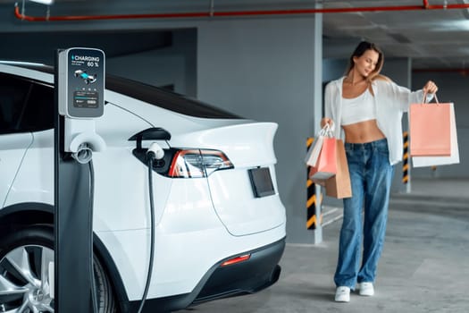 Young woman travel with EV electric car to shopping center parking lot charging in downtown city showing urban sustainability lifestyle by green clean rechargeable energy of electric vehicle innards