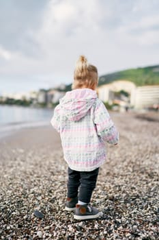 Little girl in a jacket walks along a pebbly beach, looking down at her feet. Back view. High quality photo