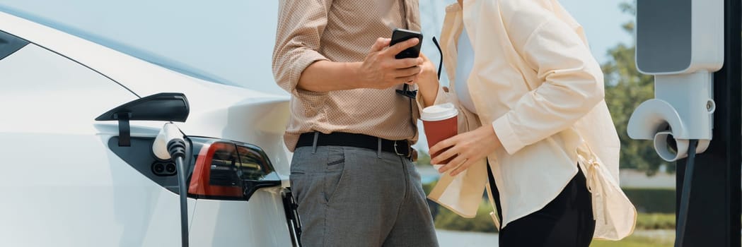 Young couple with coffee cup, recharge electric car's battery from EV charging station in green city park. Sustainable and eco friendly EV car with urban and shopping lifestyle. Panorama Expedient