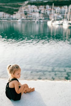 Little girl sits half-turned on the pier and looks away. Back view. High quality photo