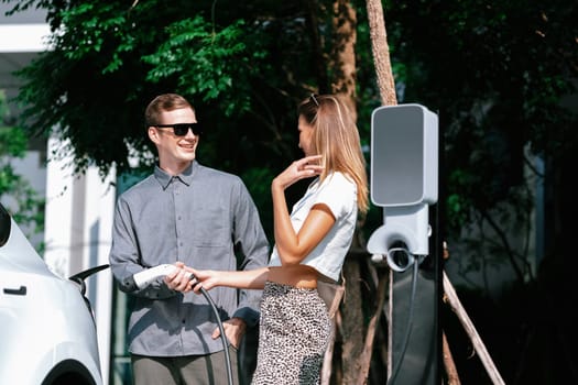 Young couple travel with EV electric car charging in green sustainable city outdoor garden in summer shows urban sustainability lifestyle by green clean rechargeable energy of electric vehicle innards