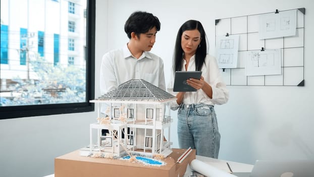 Beautiful young caucasian project manager tells asian engineer about building detail by using tablet while engineer measuring house model on meeting table with blueprint scatter around. Immaculate