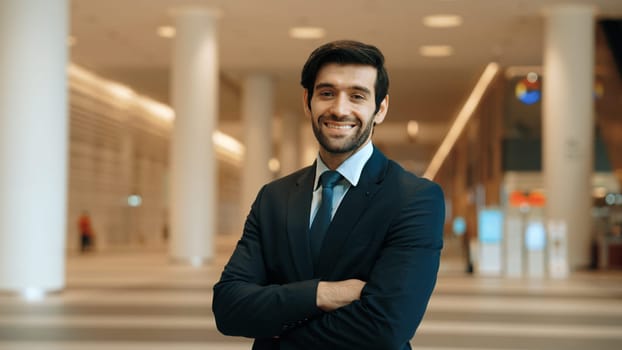 Professional business man standing with arm folded at modern mall. Smiling caucasian investor looking at camera with confident while crossing arms at hall with blurring background. Portrait. Exultant.