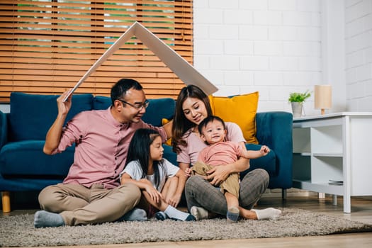 A joyful family with kids mother father sits on a sofa holding a cardboard roof symbolizing protection in their new house during relocation. Family support and planning is their path to happiness.