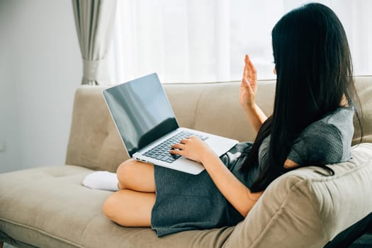 Happy woman on sofa uses laptop for video conferencing waving hi. Engaged in distant meeting discussing and smiling. Modern online communication and connection.