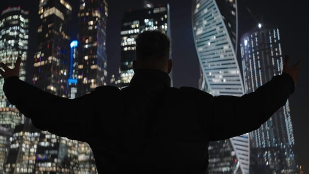 Guy raising hands on background of city. Action. Man solemnly raises hands on background of night city. Man raised hands on background of modern high-rises at night.
