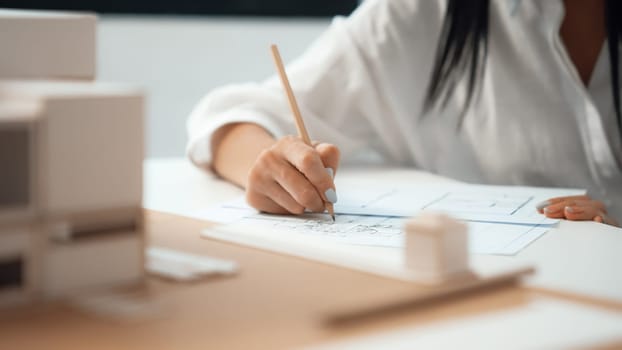 Closeup of beautiful young architect engineer hand drawing, drafting blueprint with blueprint and house model placed on working table at modern office. Focus on hand. Blurring background. Immaculate.