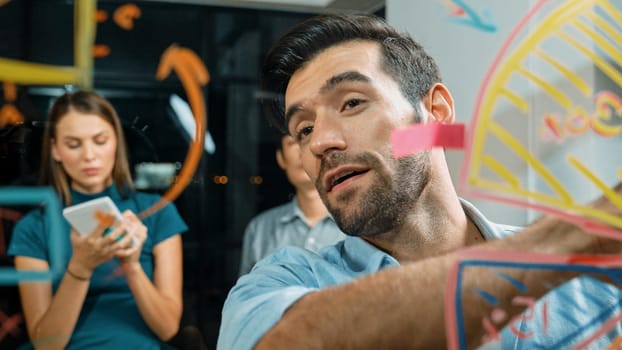 Skilled male leader explain statistic graph by drawing and writing on glass wall. Smart businessman presenting, shows financial plans to expert investor at night office. Business concept. Tracery