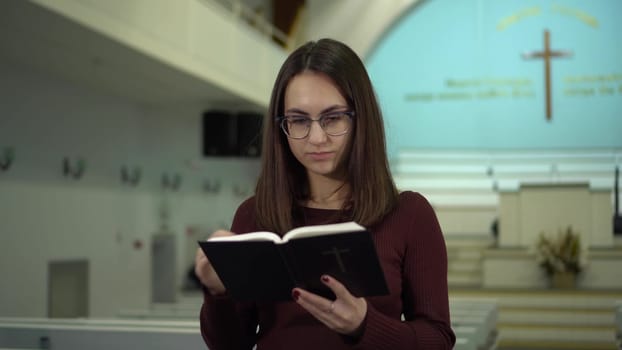 A young woman reads the Bible against the background of a cross in a church. A Protestant girl with glasses reads the Bible in church. 4k