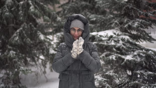 A young woman stands against a background of fir trees under heavy snowfall and rubs her hands against the cold. A girl in glasses and a down jacket with a hood stands and looks at the camera. 4k