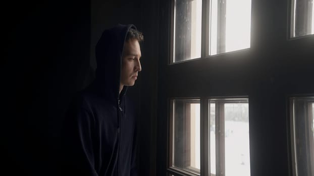 A thoughtful man at the window. Action. A young man in a dark jacket looks out of a bright window with a thoughtful face. High quality 4k footage