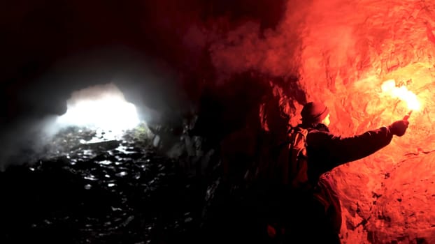 Lonely man with a red burning signal flare in a dark cavern looking for the hole, tourism concept. Male exploring underground cave.