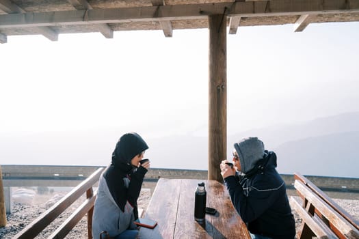 Couple of travelers sit at a table in the mountains and drink coffee with mugs from a thermos. High quality photo