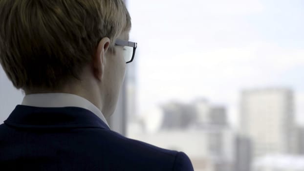 Back view of successful businessman in suit in the office looking out of the window and touching his glasses, close up. Rear view of a man looking through the window at the city