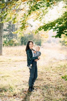 Mom with a little daughter in her arms stands in a sunny meadow near a tree. High quality photo