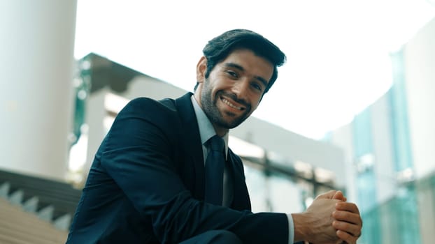 Smiling skilled businessman looking at camera while sitting on stairs. Young professional project manager smile at camera while holding mobile phone at outdoor with blurred background. Exultant.