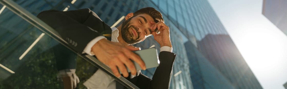 Bottom view of handsome entrepreneur using mobile phone standing on background of skyscrapers