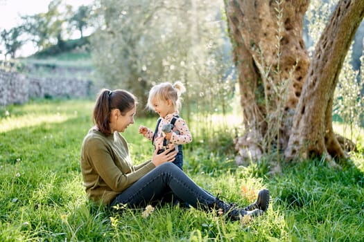 Little smiling girl stands near her mother sitting on the green grass near the tree. High quality photo