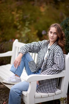 Beautiful teen blonde girl sits on a bench in an autumn park . attractive young smiling girl sitting on a bench in an autumn park. Fall Fashion Serenity: Casual Elegance in the Park