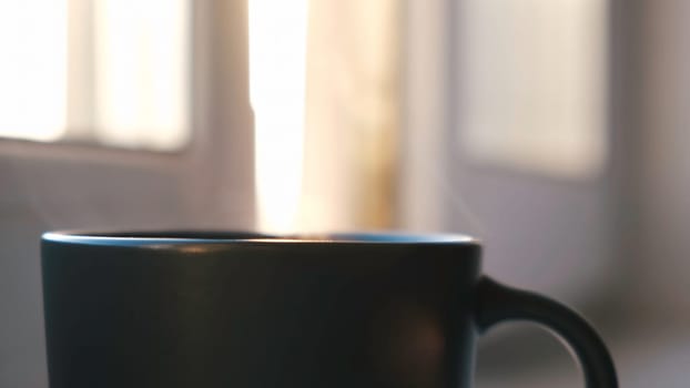 Coffee or tea mug on a window sill. Close up of the hot beverage in the dark mug with the steam rising up and the sunlight shining through the window on the background.