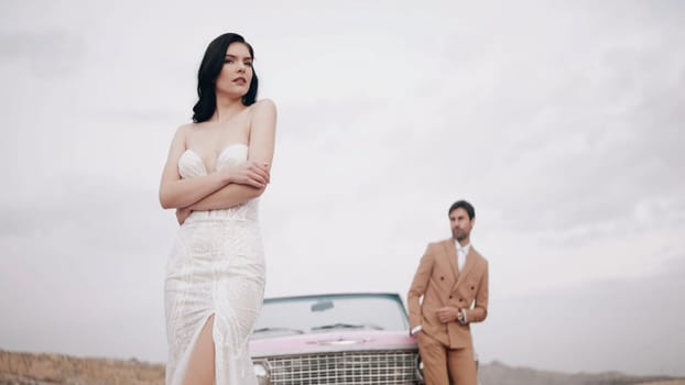 Elegant wedding photo shoot. Action. A beautiful bride in a wedding dress with a groom in a brown suit posing for a photographer on camera with happy faces in the desert next to a pink small car.