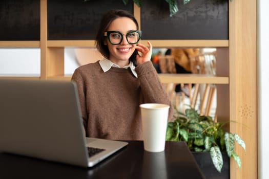 Young adult smiling woman with glasses sits at workstation. Woman poses for photo at work laptop, dressed in smart casual and glasses. Business woman sits at desk with laptop and cup of coffee