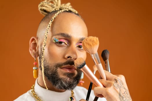 Latino gay make up artist holding makeup brush. Handsome guy wear bright make-up on isolated orange background. Fashion lgbt concept. Handsome makeup artist close up portrait