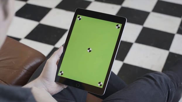 Man in dark jeans sitting on sofa and holding tablet computer with green screen on black and white checkered floor background. Ipad with chroma key.