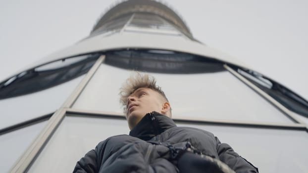 Bottom view of young man on background of tower. Action. Stylish young man in jacket at modern tower. Young man looks around warily.