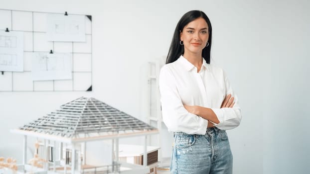 Close up portrait of young beautiful professional architect engineer cross arms and poses confidently with house model at white modern office with blueprint. Creative living and design. Immaculate.