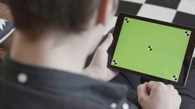 Technology and internet concept, over the shoulder view of a man tapping on the green screen of a tablet with tracking marks. Close-up of man in black shirt using his device with chroma key.