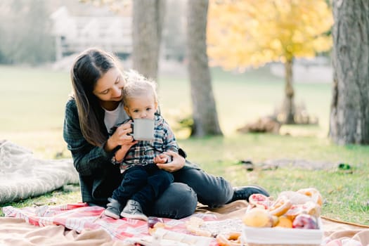 Mom gives drink to a little girl from a mug sitting on a blanket in the park. High quality photo