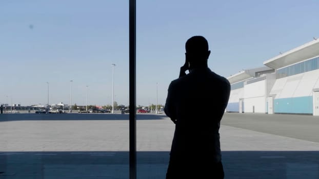 A silhouette of a businessman using smartphone, having phone call and walking in front of panoramic windows. HDR. Serious man with a smartphone indoors by the window on blue sky background