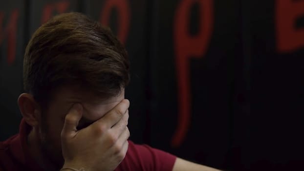 Portrait of a stressed young man covering his face with a palm against black wall with red symbols. Man making the facepalm gesture on dark wall background