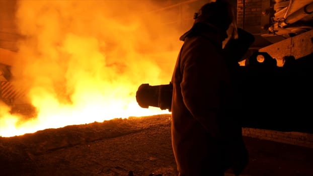 Man worker in special protective clothing standing near molten steel with sparkles and steam, heavy industry concept. Molten metal flowing in chute at the factory