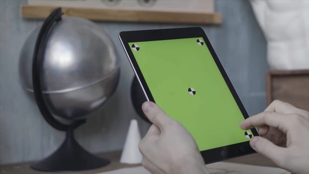 Hands of an unknown young guy tapping and scrolling on laptop with green screen on wooden table next to silver globe. Man using his Ipad with chroma key.