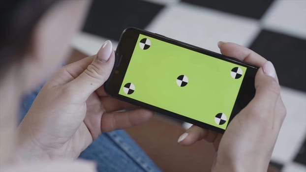 Female teen girl with dark hair using smartphone with green screen, sitting indoors. Woman holding Iphone with chroma key in horizontal position.