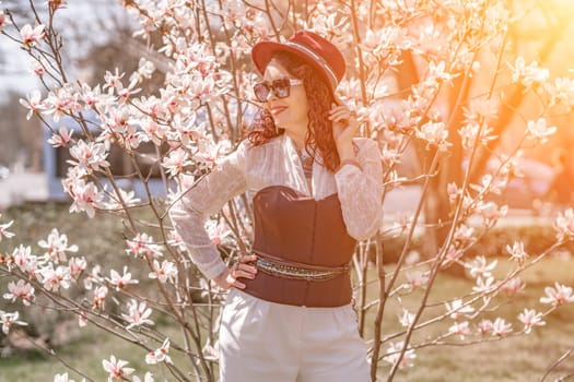 Magnolia park woman. Stylish woman in a hat stands near the magnolia bush in the park. Dressed in white corset pants and posing for the camera